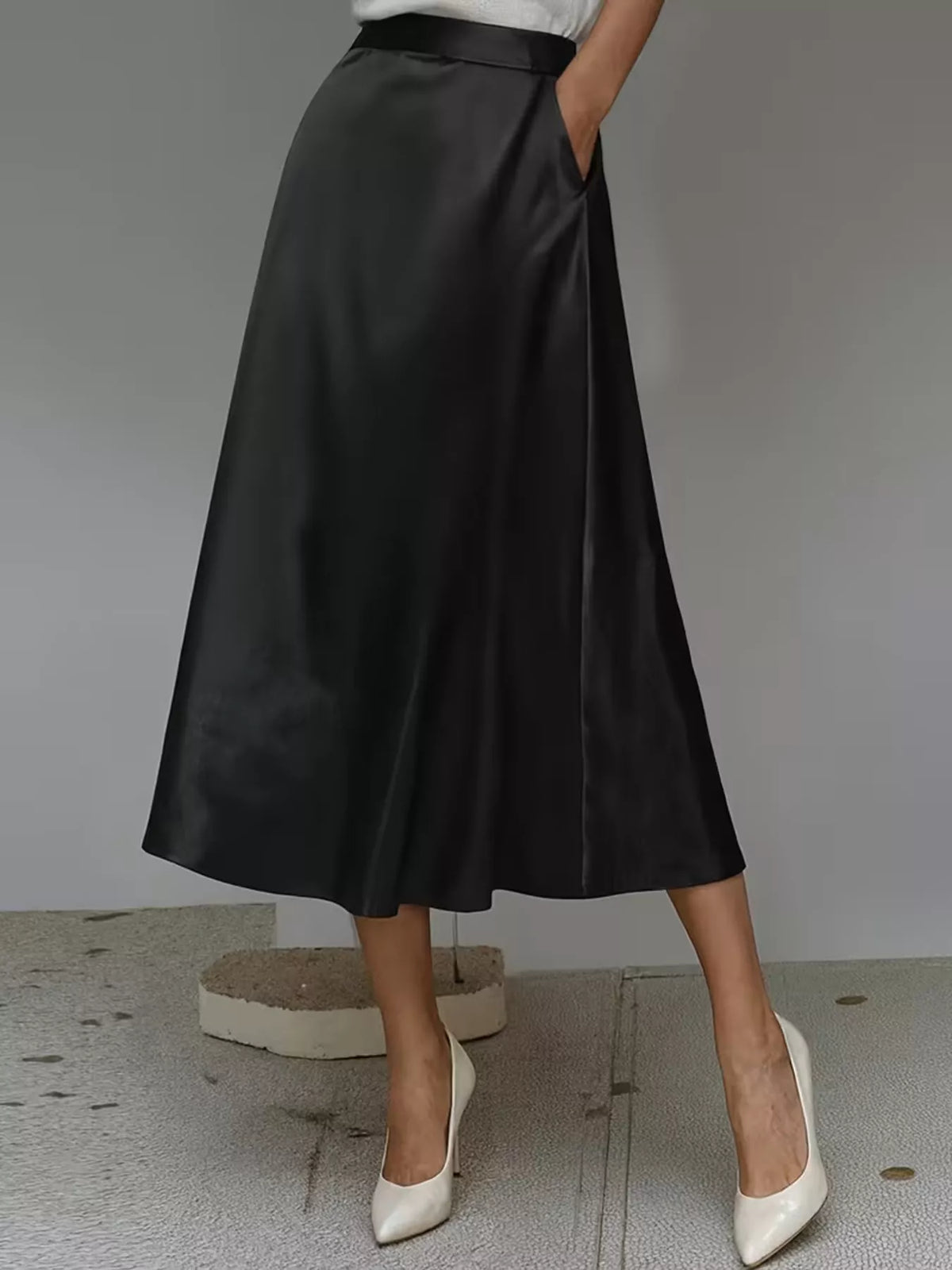 Black satin midi skirt shown at an angle, with a flowing hem and side pocket.